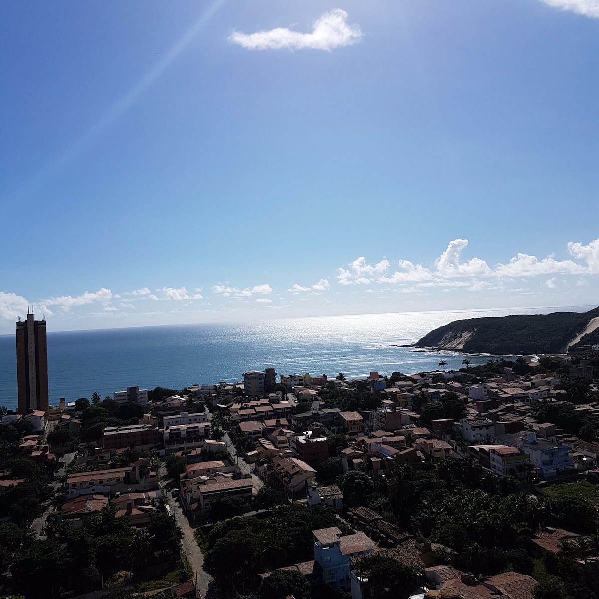 A panoramic view captures the coastline of Ponta Negra, framed by the ocean under a clear blue sky. The shoreline reflects sunlight, highlighting the sandy beach. In the foreground, a blend of residential buildings and greenery creates a serene landscape.