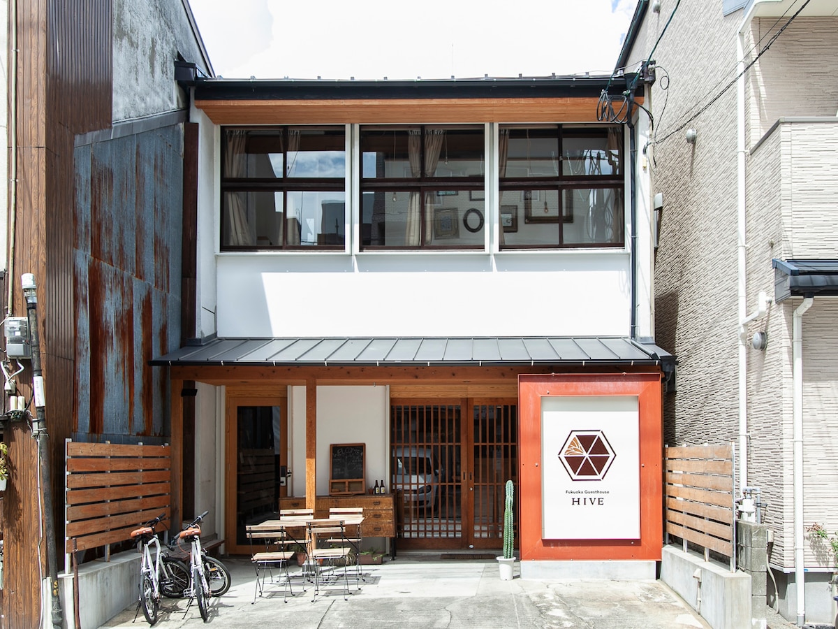 The exterior of the guesthouse features a modern design with a contrasting combination of wood and metal elements. Large windows allow natural light to enter the space, while a vibrant sign displays the name 'HIVE' prominently. Outdoor seating is available near the entrance.