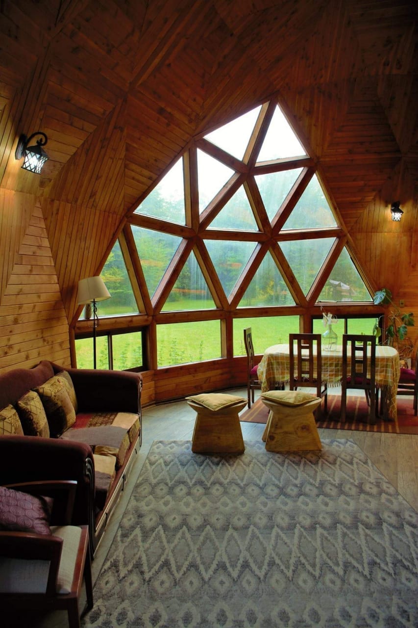 A cozy interior is highlighted by a geometric wooden design and large windows that offer views of the surrounding nature. A seating area includes a sofa and two wooden stools. A dining table is positioned near the window, illuminated by natural light.