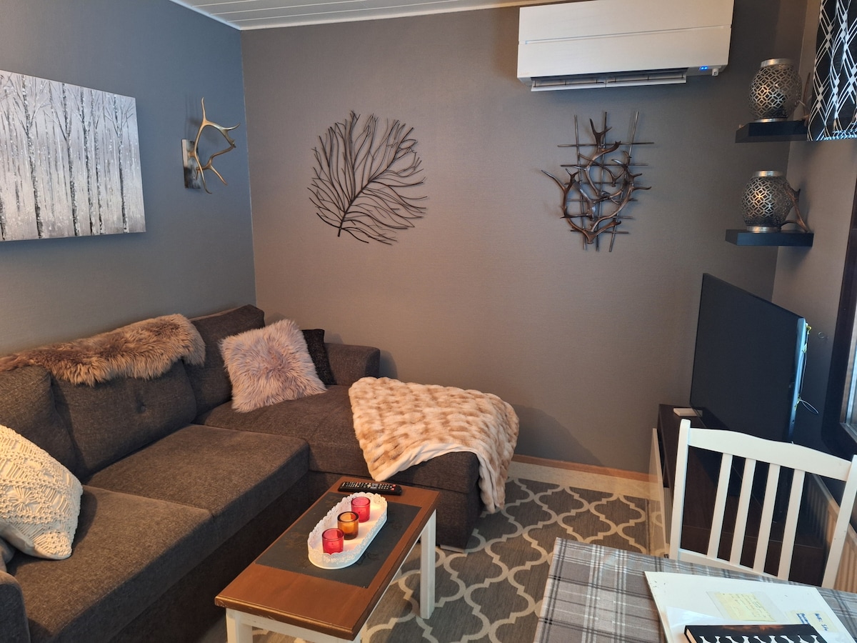 The cozy living area features a dark grey sectional sofa adorned with textured cushions and a soft throw. Wall art displaying natural elements adds character, while shelves showcase decorative pieces. A small dining table is positioned nearby, with a centerpiece of candles providing a warm touch.