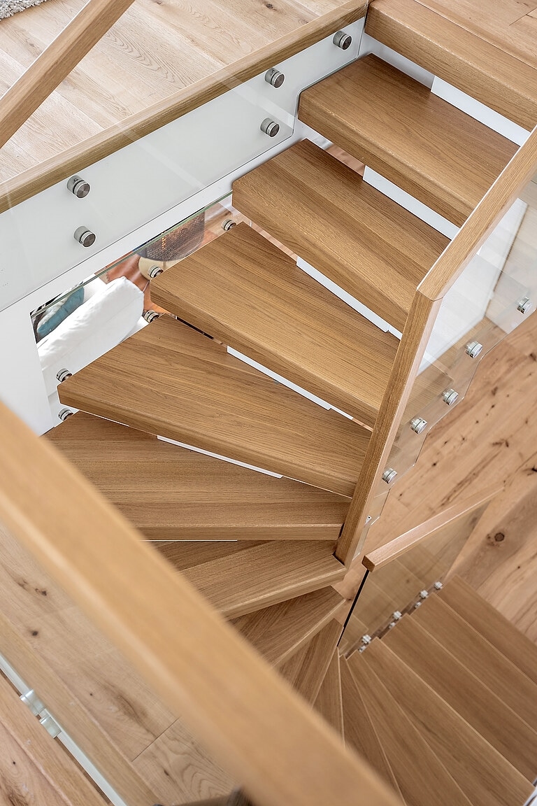 A spiral staircase features elegant wooden steps leading to an upper level. Glass panels provide safety and transparency, allowing for an unobstructed view of the surrounding interior. The warm tones of the wood complement the flooring, creating a cohesive design in the space.