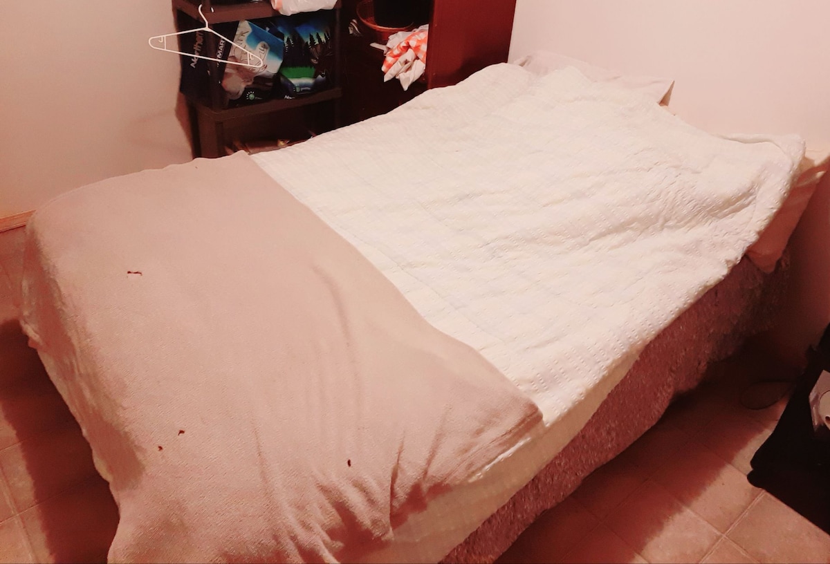 A spacious bed is centered in the room, covered by a light-colored quilt and a warm-toned blanket. Nearby storage shelves hold various items, and a soft carpeted floor is visible. The room has a neutral color palette, contributing to a relaxed atmosphere.