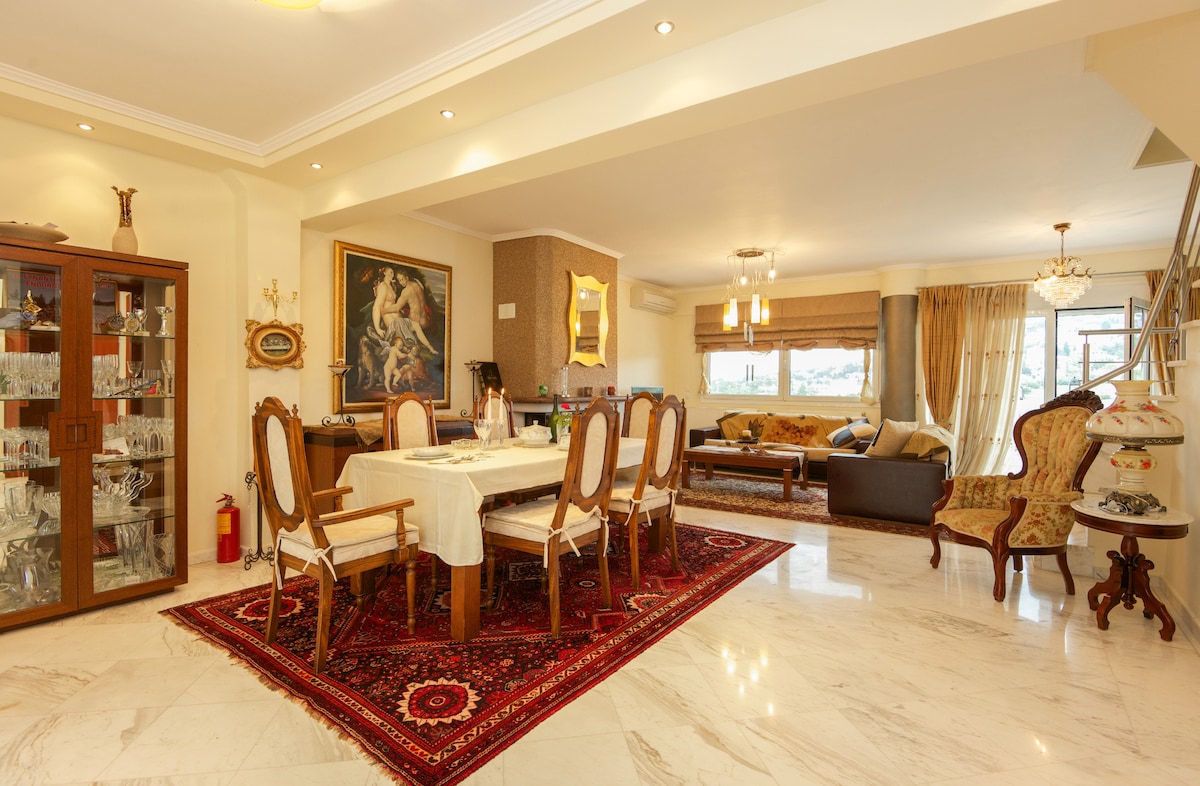 A spacious dining area displays a wooden table surrounded by six elegant chairs. A glass-fronted cabinet features an array of glassware. The living area is visible in the background, showcasing a comfortable seating arrangement and large windows allowing natural light.