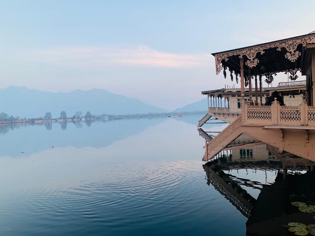 A houseboat is pictured reflecting in the calm waters of Nigeen Lake, surrounded by distant mountains. Soft pastel hues are visible in the sky, creating a serene environment. The architectural details of the houseboat's wooden structure are highlighted, accentuated by traditional carvings.