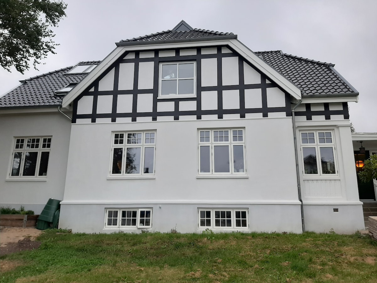 The exterior of a two-story house features a mix of white and black paneling, with multiple windows allowing for natural light. A well-maintained grassy area surrounds the base, and a small garden area is visible to the left of the image.