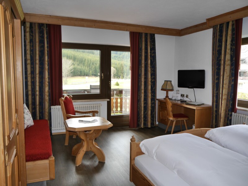 A bright room features a spacious bed, wooden furniture, and a cozy seating area with a red chair. Two windows provide natural light and views of nature. A flat-screen television and a work desk are positioned against the wall, enhancing the functional layout.