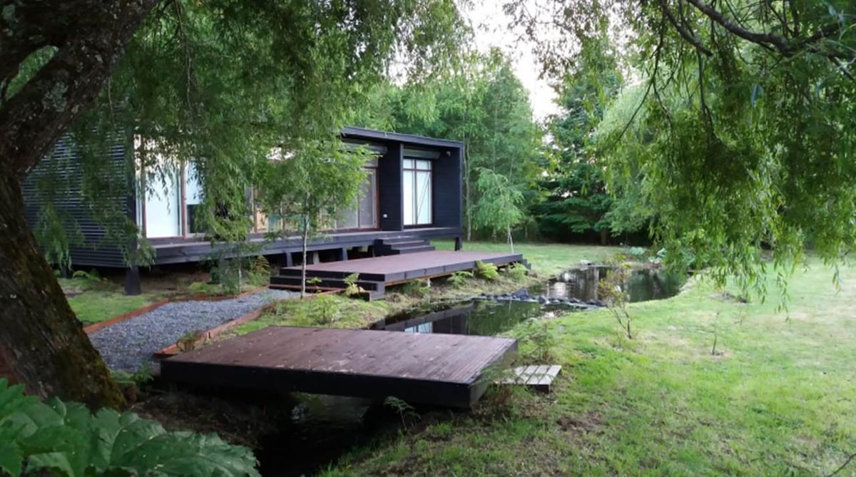 The exterior of a modern house is depicted, nestled within a lush, green garden. A wooden deck extends toward a tranquil stream, surrounded by neatly maintained grass. Tall trees provide shade, enhancing the serene atmosphere of the natural setting.