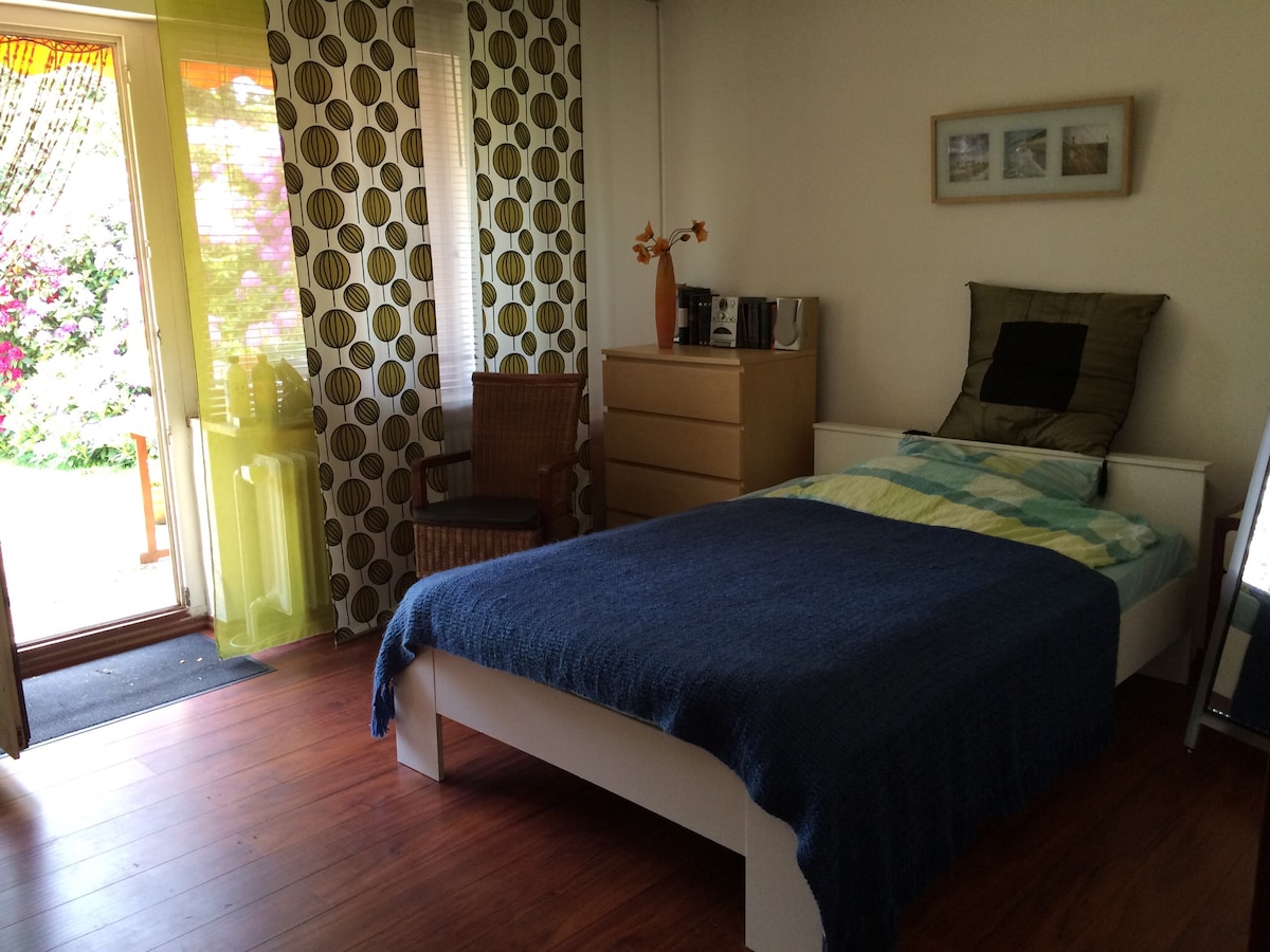 A bedroom features a single bed draped with a blue blanket, accompanied by a wooden dresser and a chair. Natural light enters through a door leading to a garden, enhanced by patterned curtains. A small table is visible beside the bed, creating a cozy atmosphere.