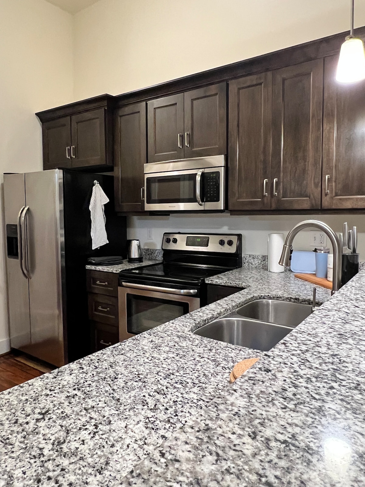 A modern kitchen features dark wooden cabinetry and sleek stainless steel appliances. A spacious granite countertop includes a double sink and a stove, complemented by a microwave and a refrigerator. Soft lighting highlights the layout and reflects on the polished surfaces.
