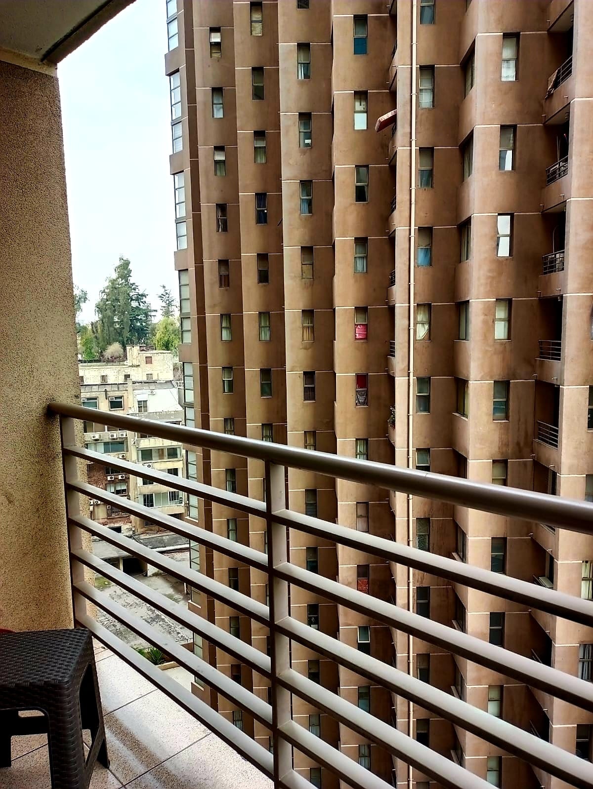 A balcony area is visible, featuring a metal railing and a small table and chair. The view includes a neighboring residential building with a textured brown facade and multiple windows. A glimpse of surrounding rooftops and trees can also be seen in the background.