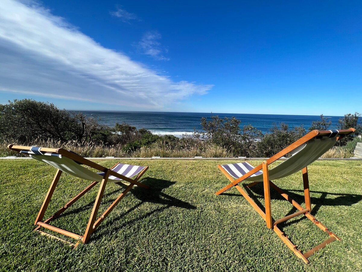 Two wooden deck chairs with striped fabric are positioned on a green lawn, facing the ocean. The blue sky and sea create a serene backdrop, while the shoreline is visible with gentle waves lapping against the shore.