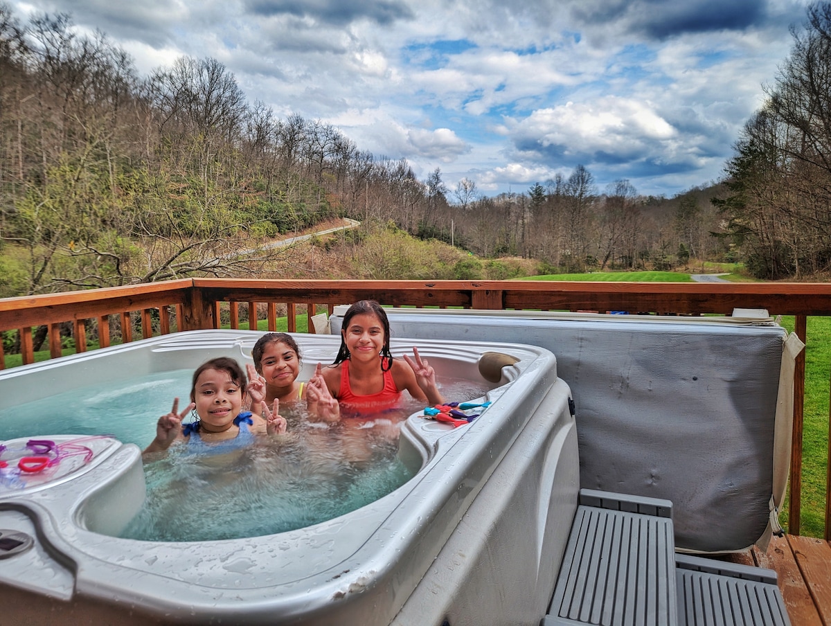A hot tub is set on a wooden deck, surrounded by lush greenery and rolling hills. Three individuals relax in the tub with bright swimsuits, enjoying the serene outdoors under a cloudy sky. This area provides a peaceful retreat for unwinding after day activities.