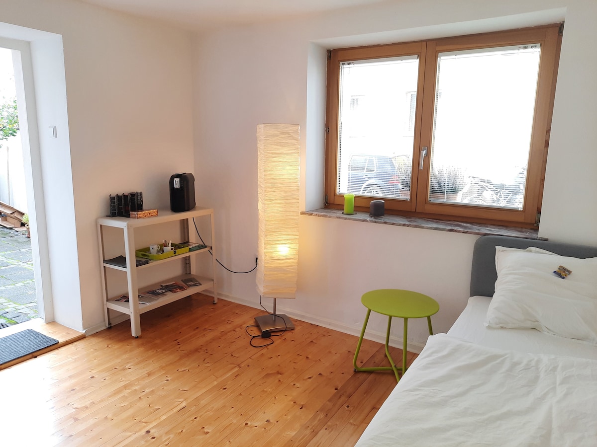 A bright room features a cozy bed with crisp linens and natural light streaming through a large window. A standing lamp provides soft illumination next to a small green table. A shelf displays miscellaneous items, complemented by a coffee machine nearby.