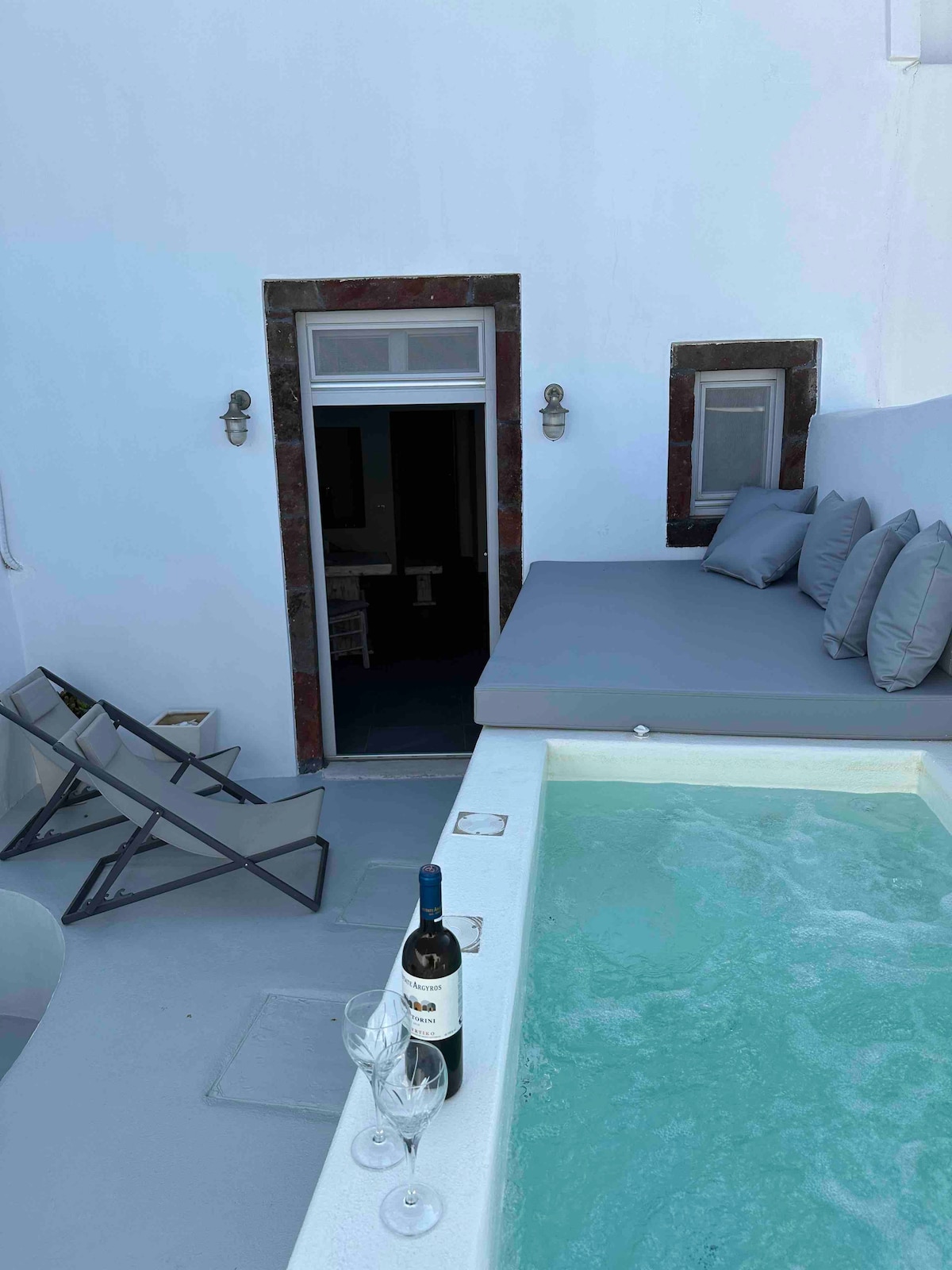 An outdoor area features a hot tub with clear water, accompanied by two wine glasses and a bottle of wine nearby. A spacious seating area is adorned with gray cushions, and two folding chairs are positioned on the side, providing additional relaxation options.