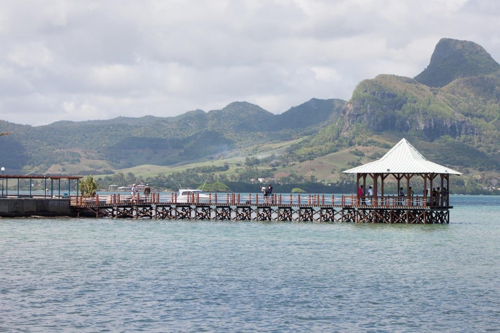 A wooden pier extends over calm waters, leading to a pavilion with a pointed roof. Lush green hills and mountains serve as a backdrop, showcasing the natural beauty of the area. A few figures can be seen enjoying the view from the pier.