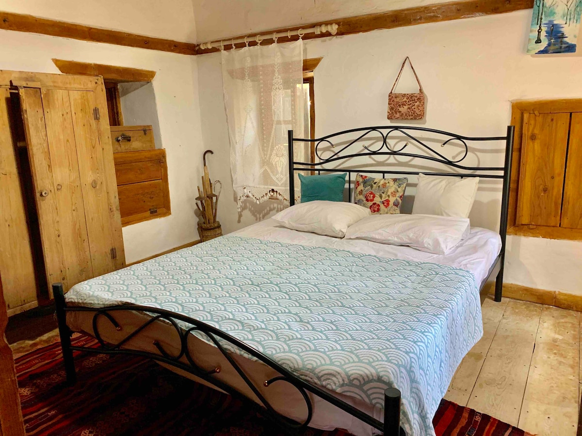 A charming bedroom features a black metal bed with a patterned blanket and two white pillows. Wooden doors are positioned on opposite walls, and a small textile wall hanging adds a decorative touch. Soft light filters through the lace curtain, creating a calm ambiance.