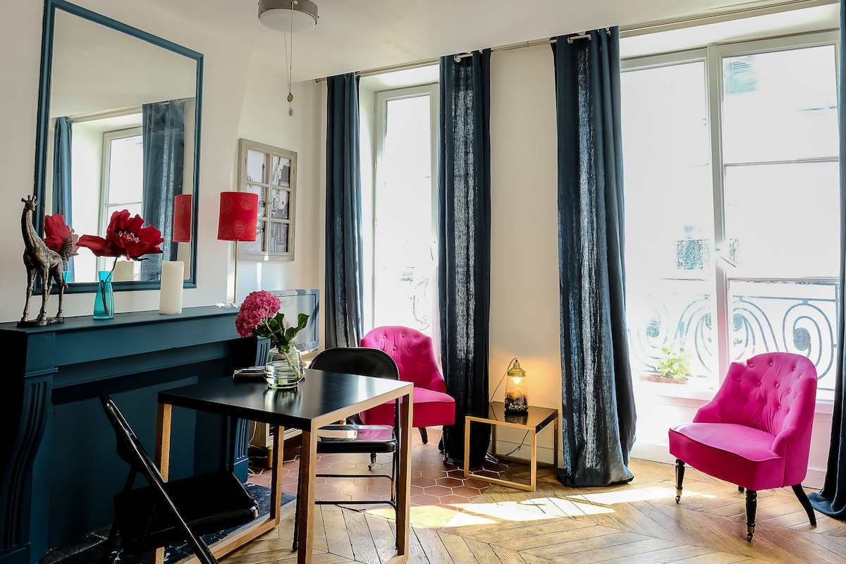 The living area features two pink chairs flanking a small table, with a decorative lamp beside it. A tall mirror reflects light and space. Large windows are draped with dark curtains, allowing natural light to illuminate the wooden floor, showcasing its varied textures.