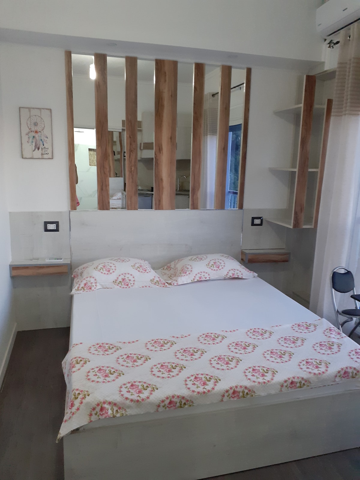A bright sleeping area features a spacious bed with a floral-patterned blanket. Wooden paneling adds texture to the room, while built-in shelves provide storage space. A small chair is positioned nearby, and natural light is allowed in through a window.