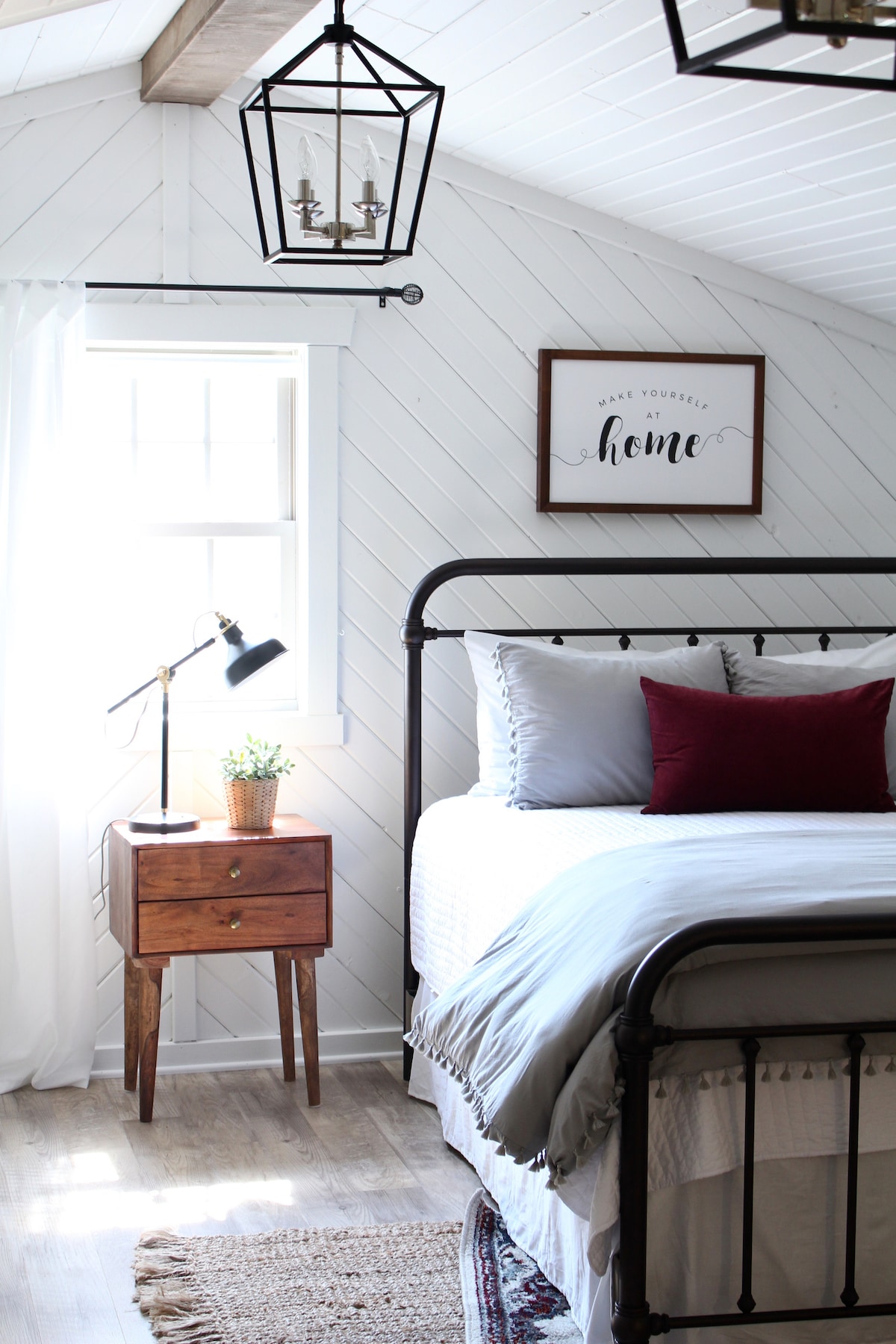 A bright sleeping area features a metal bed with plush bedding and a decorative pillow. Beside the bed, a wooden nightstand holds a small potted plant. A modern lamp provides soft lighting, while a framed sign with the text 'Make yourself home' decorates the wall. Natural light enters through a window, with sheer curtains gently filtering it.