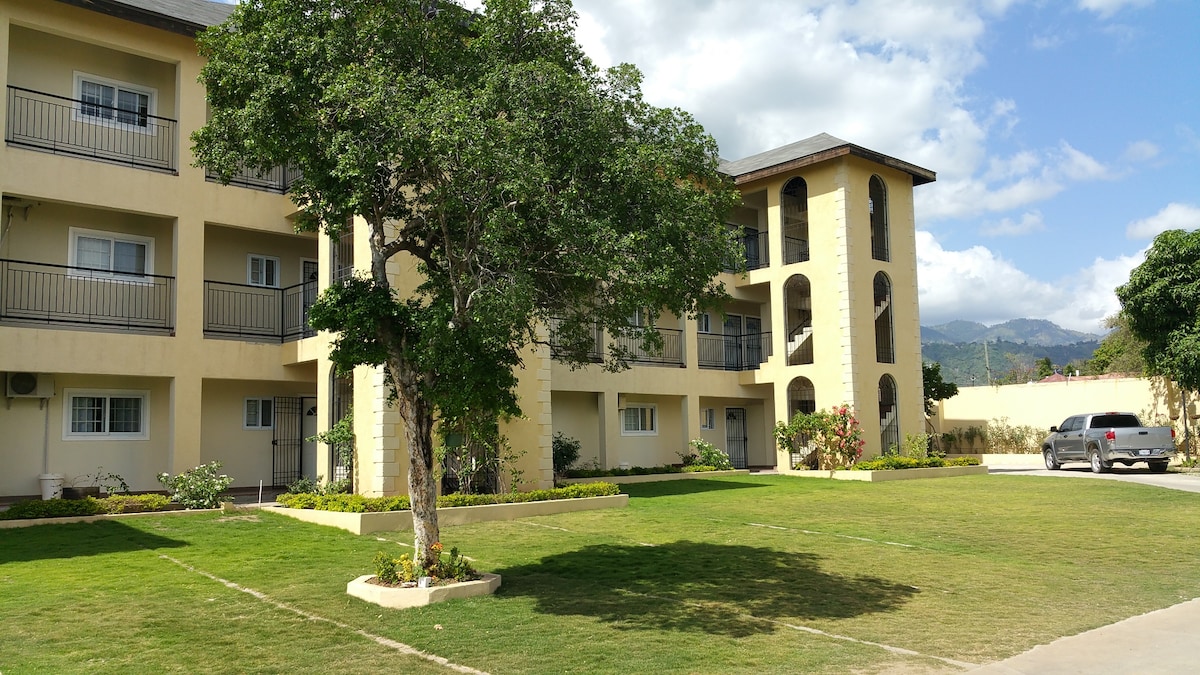 An apartment building is showcased with a light-colored exterior, featuring multiple balconies. Green grass and landscaped gardens surround the structure. A tree provides shade in the front yard, while a parked vehicle is visible in the parking area. The mountains are faintly seen in the background.