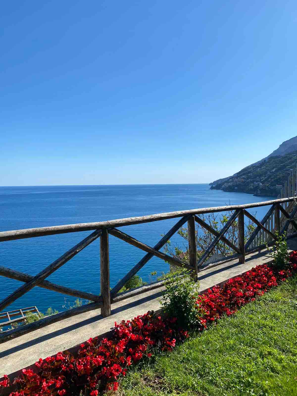 A wooden fence encloses a grassy area adorned with vibrant red flowers, offering a clear view of the calm blue sea under a cloudless sky. The coastline extends in the distance, highlighting the natural beauty of the Amalfi Coast.