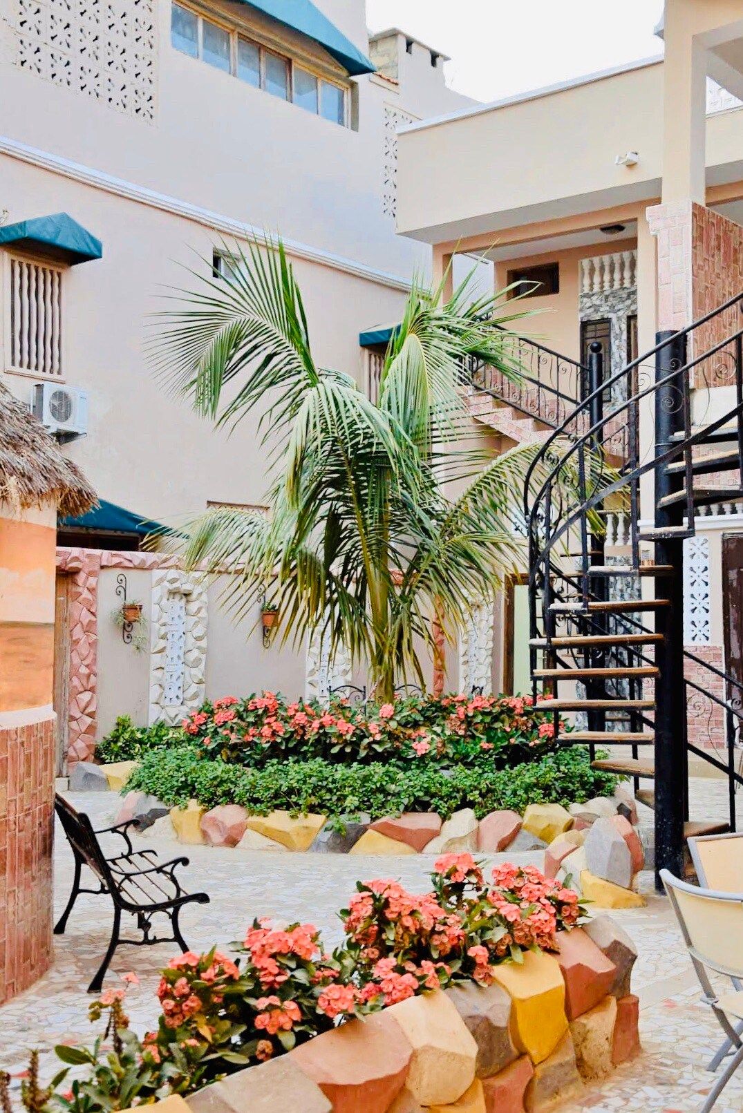 A peaceful courtyard is surrounded by colorful flower beds and a leafy palm tree. A spiral staircase leads to upper levels, while a black bench offers a spot to relax amid the lush greenery and vibrant blooms.