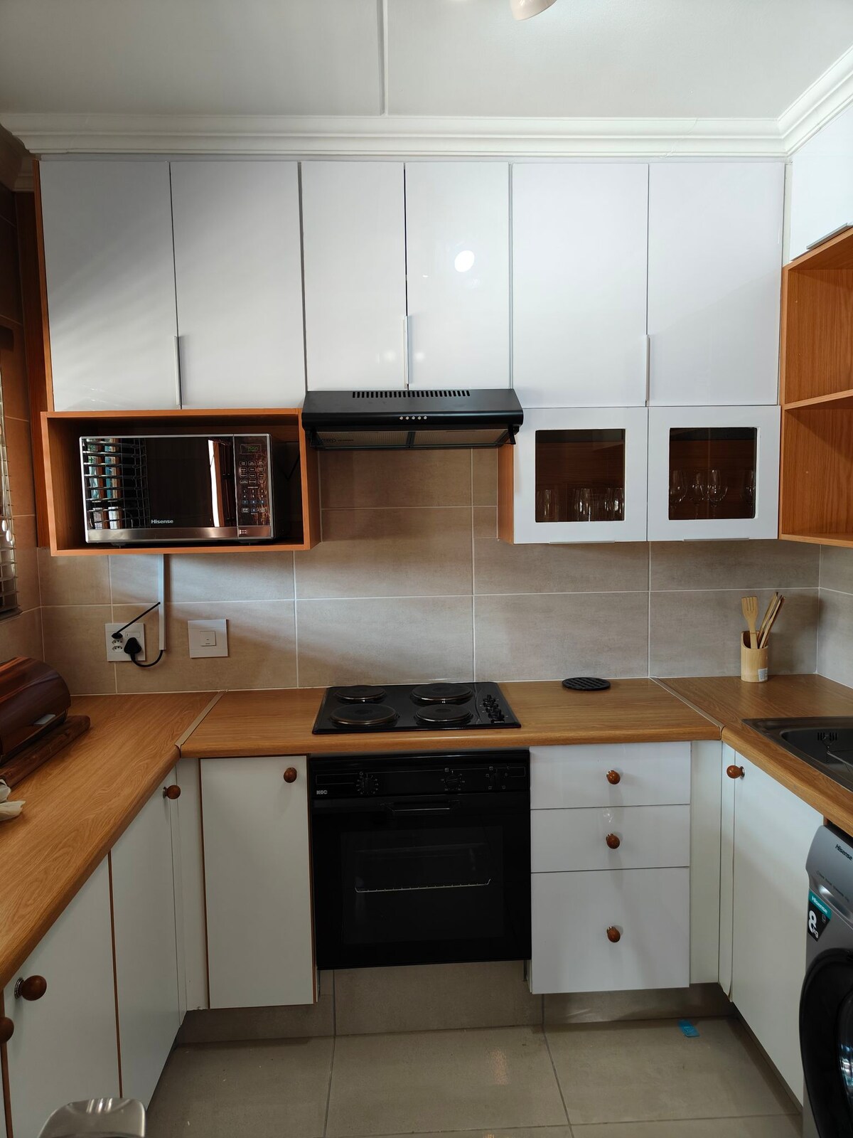 A modern kitchen is showcased with a blend of white and wood cabinetry. Appliances include a microwave, stove, and an oven, all neatly arranged. The countertop is clear, providing ample workspace, while natural light enters through a nearby window.