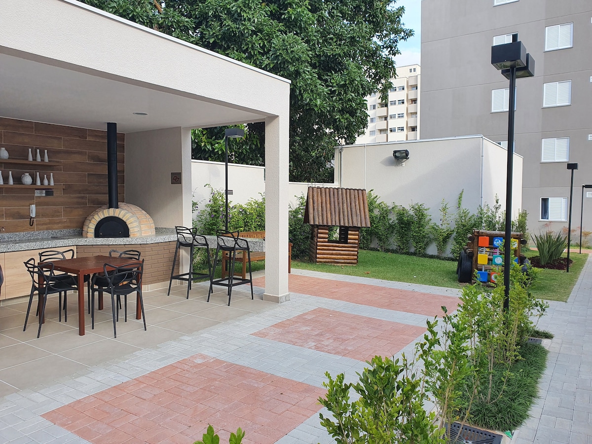 An outdoor communal area includes a shaded seating space with several chairs and a large pizza oven. A small grassy area features a playhouse, bordered by neatly arranged plants and paving stones. Modern lighting fixtures enhance the atmosphere in this inviting space.