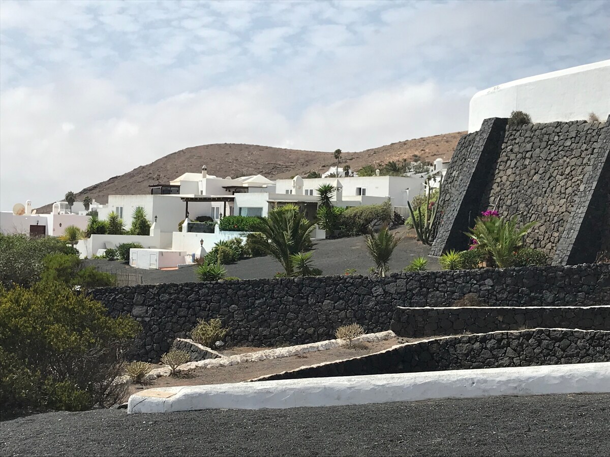 A view of the villa complex is showcased, featuring white buildings set against a backdrop of a rocky hillside and desert landscape. The lush green palm trees and vibrant plants add contrast to the natural stone pathways and dark volcanic ground.