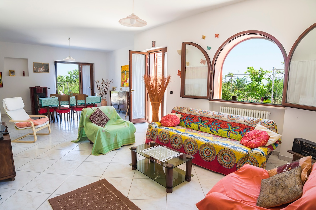 The living area features multiple seating options, including a patterned sofa and lounge chairs. Large windows allow natural light to illuminate the space, revealing views of the surrounding greenery. A dining table is set in the distance, complemented by colorful decorative elements on the walls.