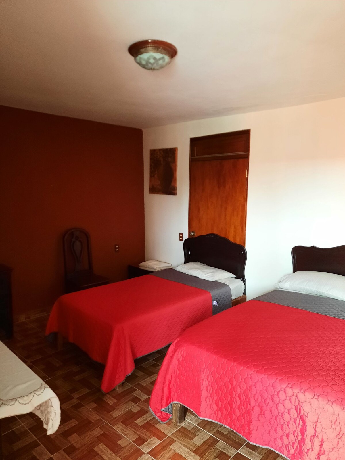 A cozy room features a double bed and a single bed, both covered with red bedspreads. A wooden wardrobe is visible along one wall, complemented by a simple chair. The space is designed with warm earth tones, providing a calm atmosphere.
