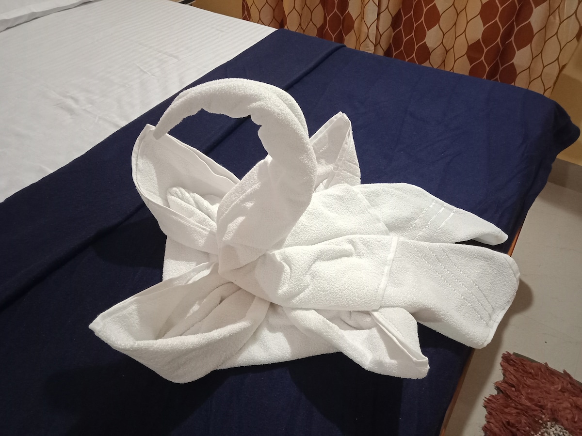 A decorative arrangement of towels is presented in the shape of a swan on the bed. The bed features crisp white linens with a navy blue throw. Soft lighting enhances the simple yet thoughtful touch, providing a welcoming appearance to the space.