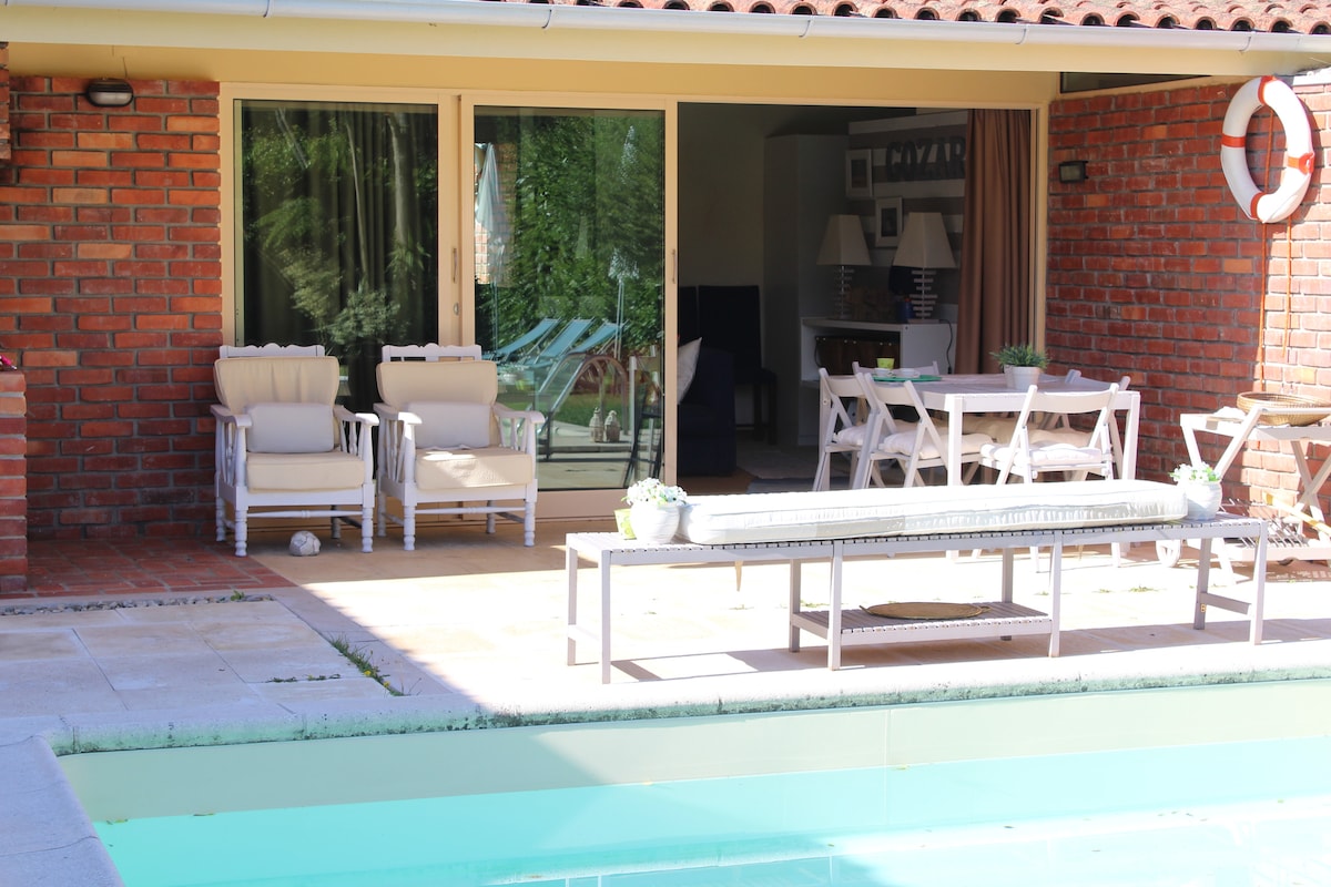 A sunlit patio area features comfortable seating options near the salt-water pool. A long, simple bench complements a nearby dining table. Sliding glass doors provide a view into the interior space, which includes light-colored furnishings and a warm ambiance.