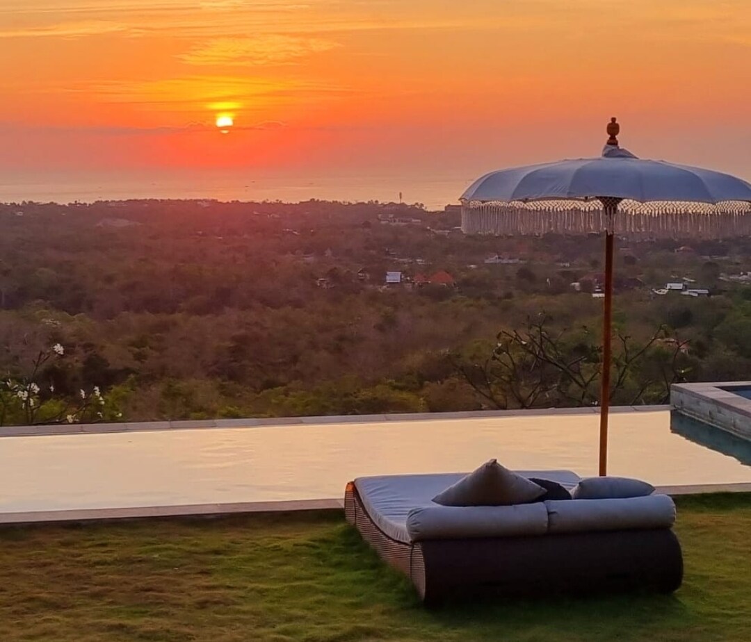 A tranquil sunset is captured over the horizon, painting the sky with warm hues of orange and pink. A sunbed sits elegantly near the infinity pool, accompanied by a parasol. Lush greenery extends towards the ocean, enhancing the serene view.