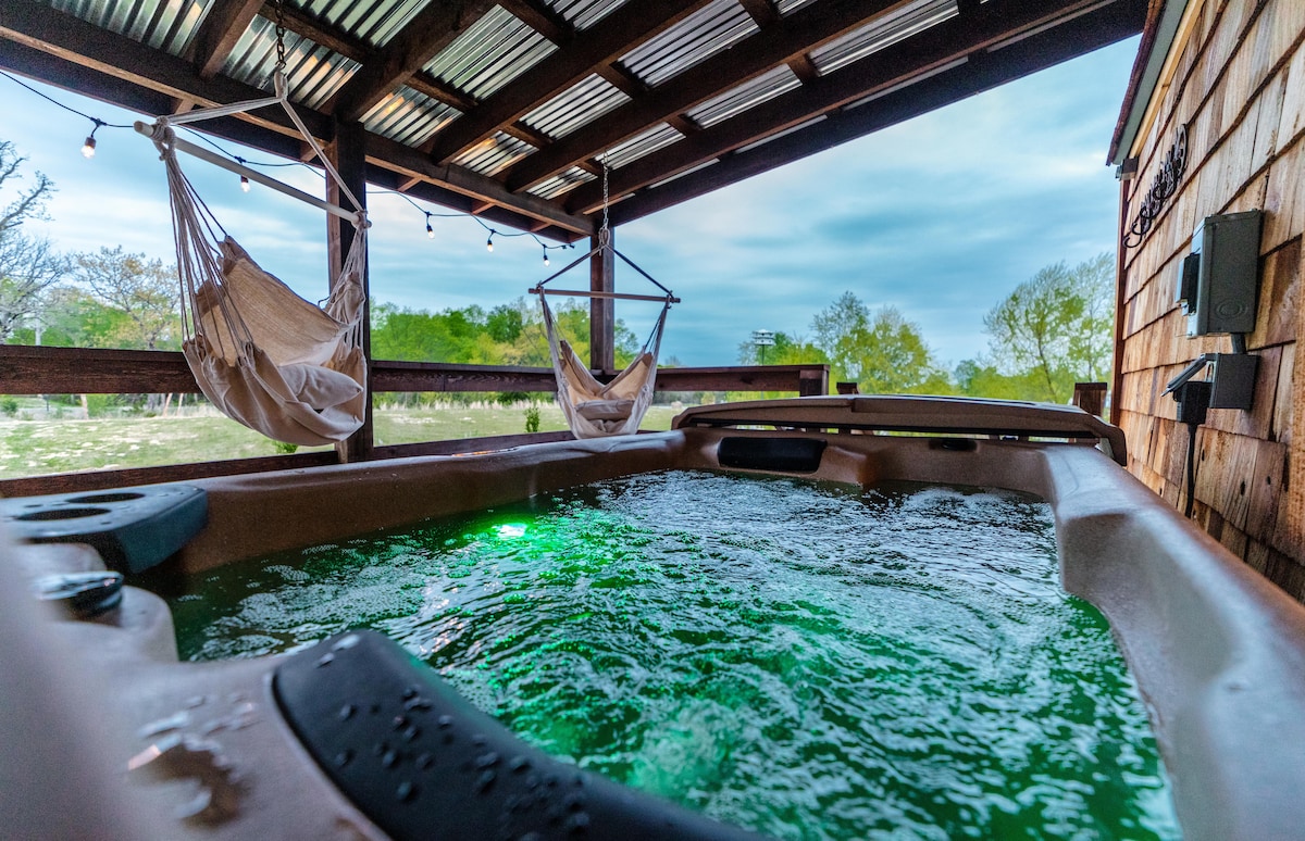 A hot tub is situated on a covered deck, featuring bubbling water illuminated in green. Two hanging chairs are positioned nearby, offering a cozy seating option. The outdoor area is surrounded by lush greenery, creating a relaxing atmosphere.