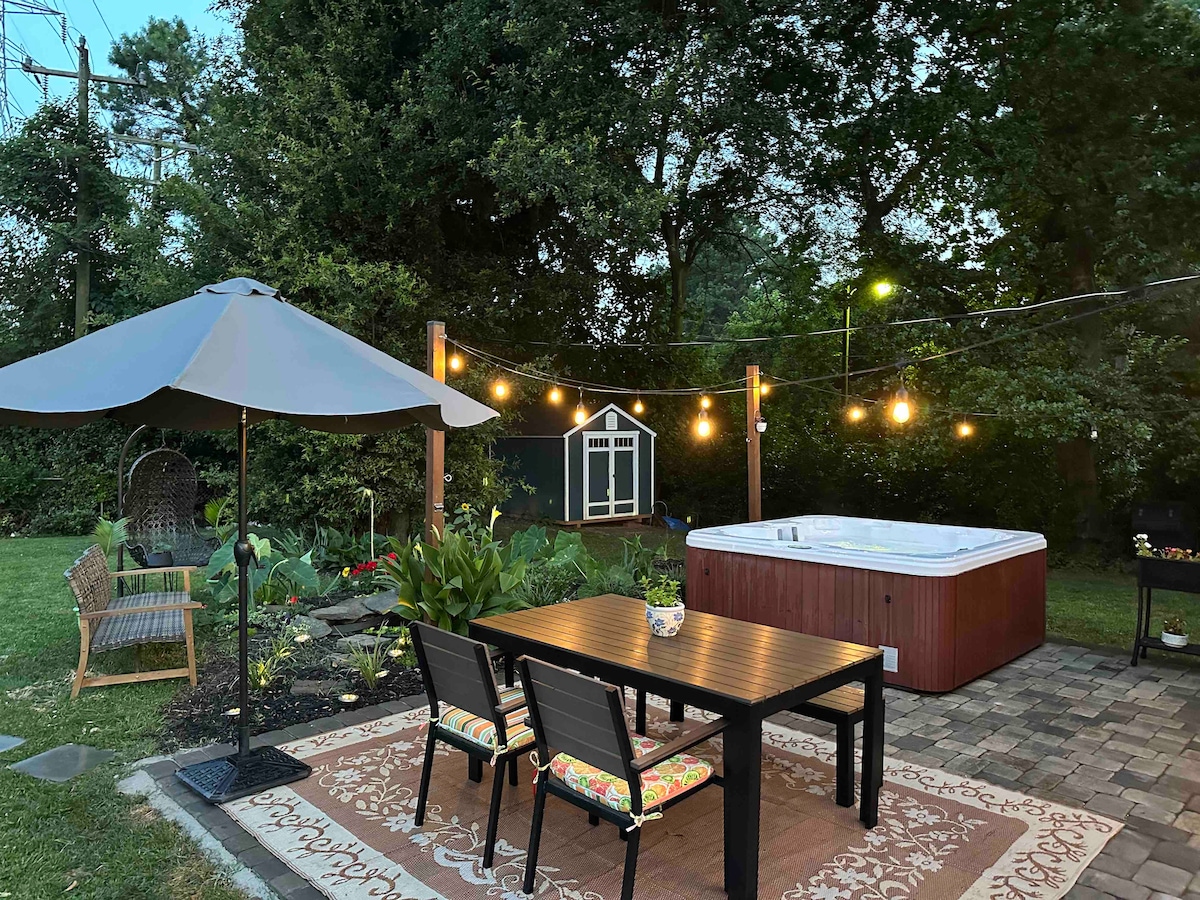 A welcoming outdoor area features a wooden table surrounded by four chairs. A large umbrella provides shade, while string lights illuminate the space. A hot tub is located nearby, and a small shed is present in the background, framed by lush greenery.