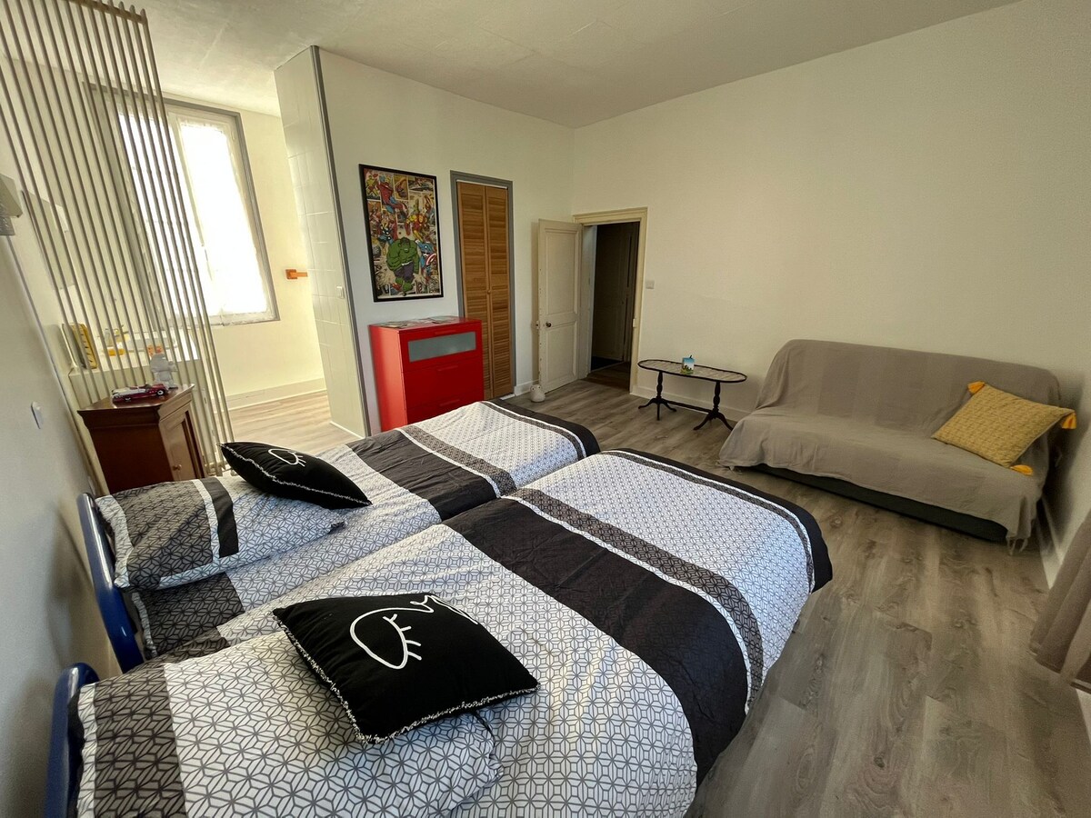A spacious room features two separate beds dressed in patterned bedding. A simple sofa is placed against the wall, complemented by a decorative pillow. Natural light enters through large windows, illuminating the neutral-toned walls and wooden floors.