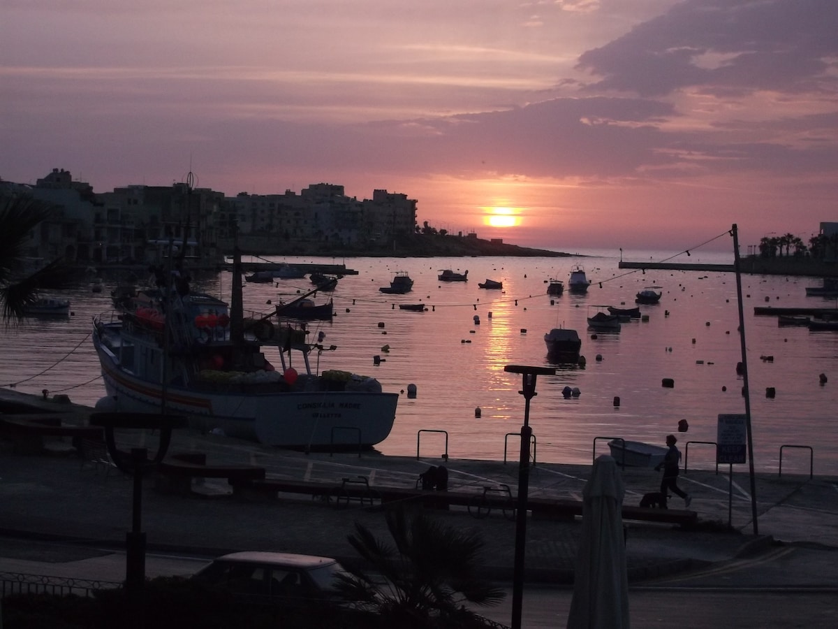 A serene waterfront scene at sunset displays a tranquil harbor filled with boats. The sky is painted in soft pinks and purples, reflecting on the water's surface. Silhouettes of distant buildings can be seen along the shore, while a figure strolls along the promenade.