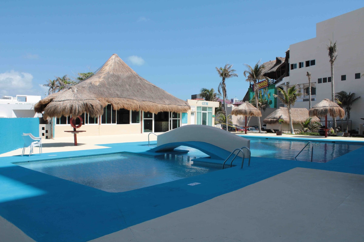 A spacious outdoor swimming pool is bordered by a bright blue surface. A thatched-roof gazebo is visible nearby, along with an archway leading into the pool. Palm trees provide a tropical ambiance, and a clear sky enhances the inviting atmosphere.