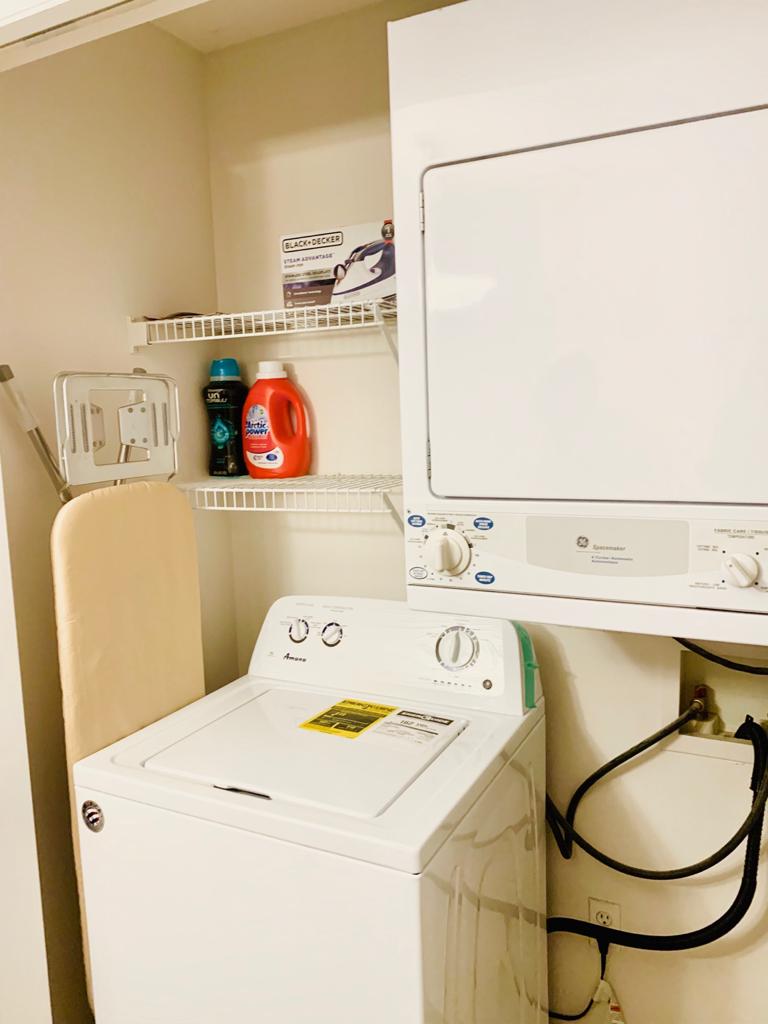 A compact laundry area is visible, featuring a stacked washer and dryer. Shelves above contain laundry supplies, including fabric softener and detergent. An ironing board is positioned nearby, with an iron resting against it for convenience.