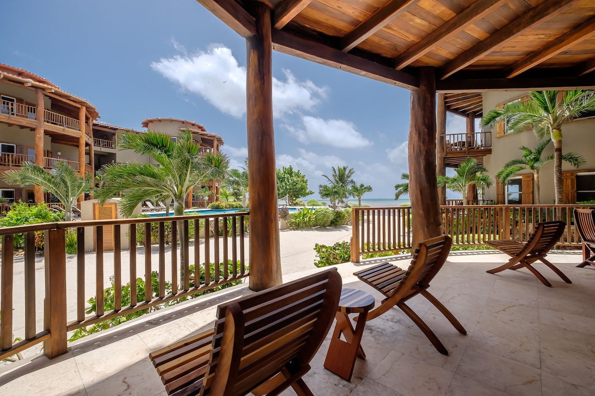 A covered patio offers a view of the lush landscaping and resort-like atmosphere. Two wooden lounge chairs are positioned facing a spacious courtyard, framed by palm trees and balconies of nearby buildings, creating a peaceful setting for relaxation.