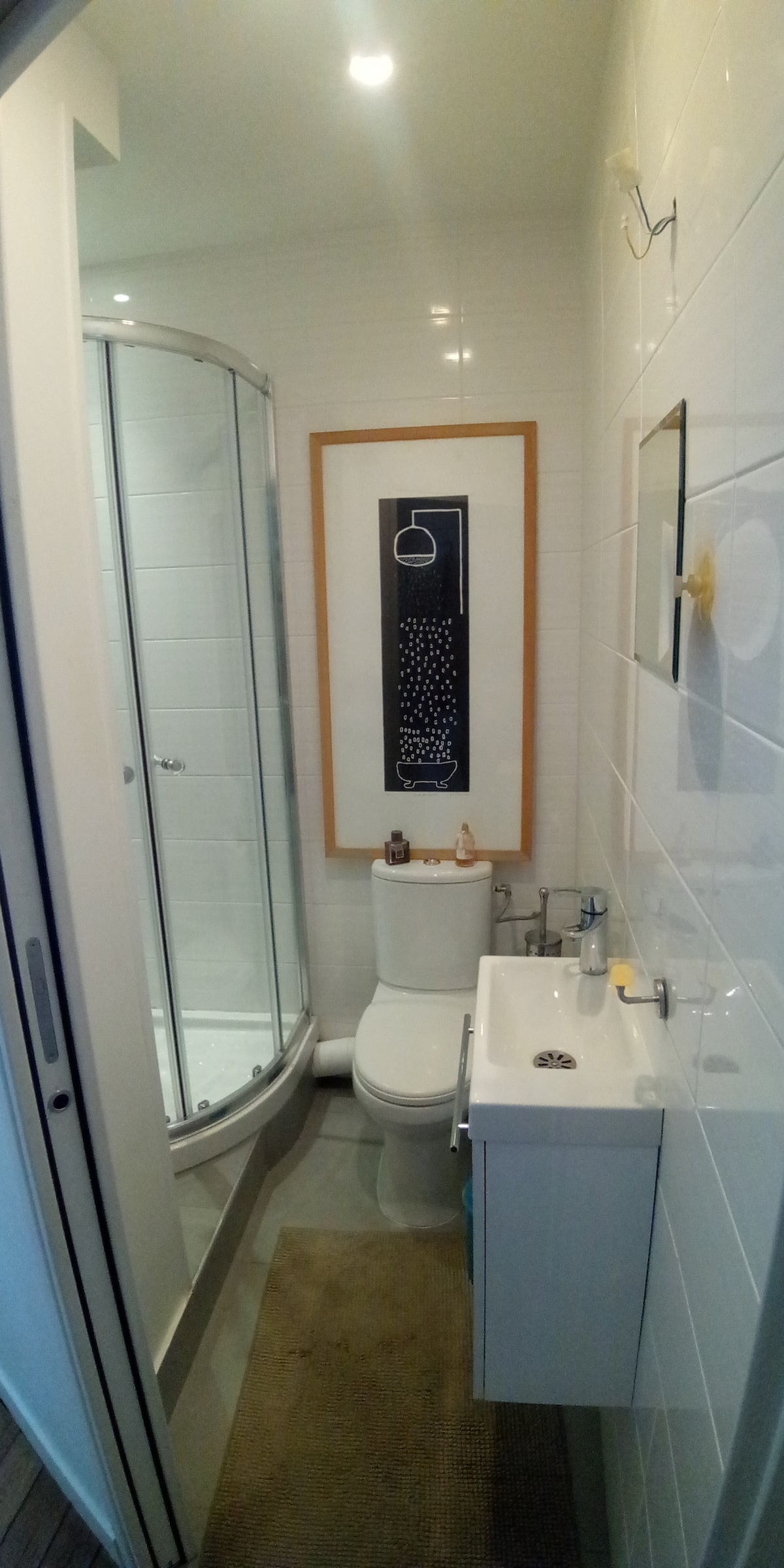 A compact bathroom is visible, featuring a glass shower enclosure and a modern toilet. A small sink with storage is mounted on the wall, and a piece of framed art is displayed above. Soft lighting enhances the clean, white tiled surfaces.