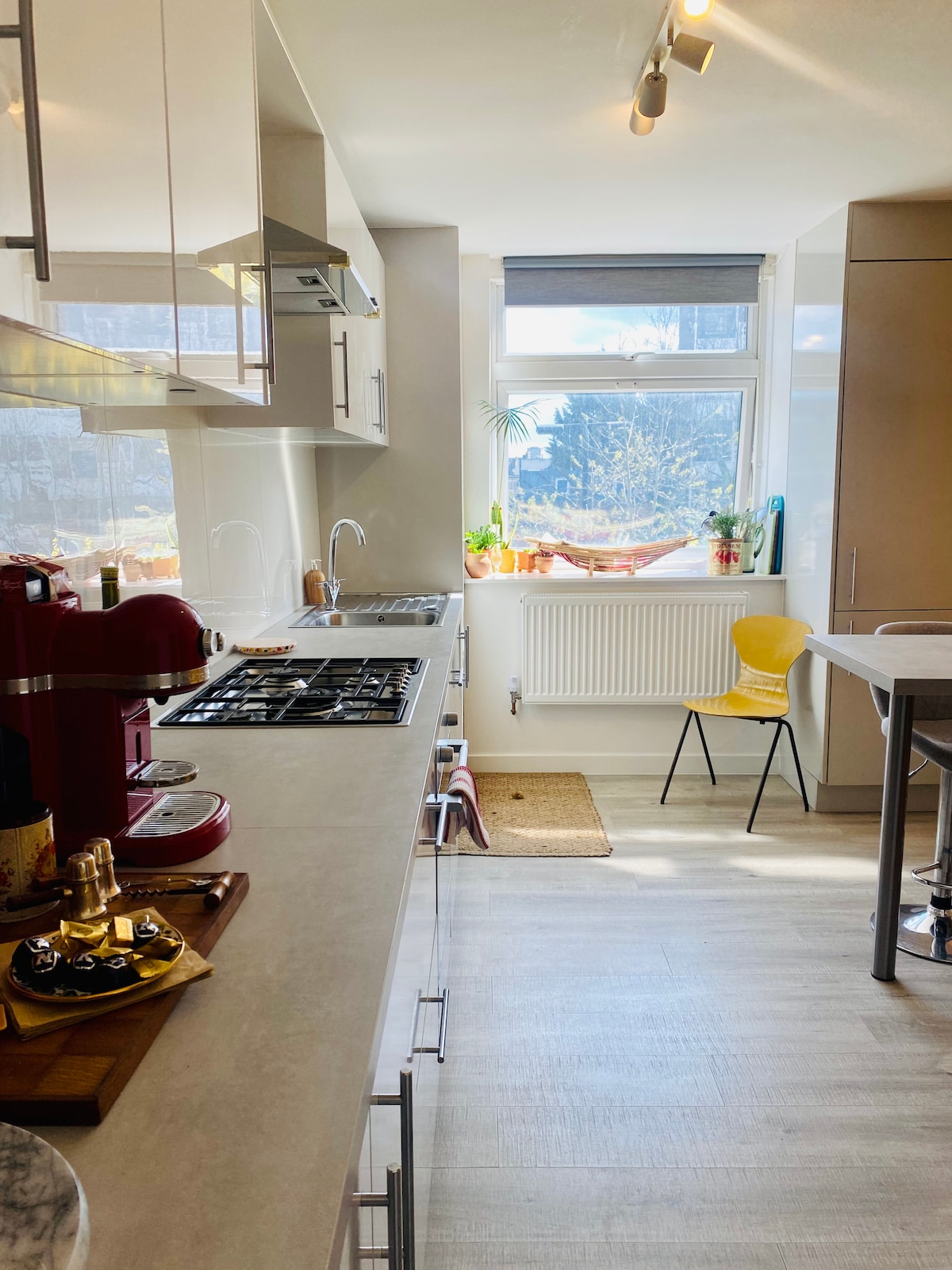 A bright kitchen features modern appliances and sleek cabinetry. A coffee machine and an array of utensils are positioned on the countertop. Ample natural light enters through a window, illuminating the space. A small dining area is visible, complemented by a cheerful yellow chair.