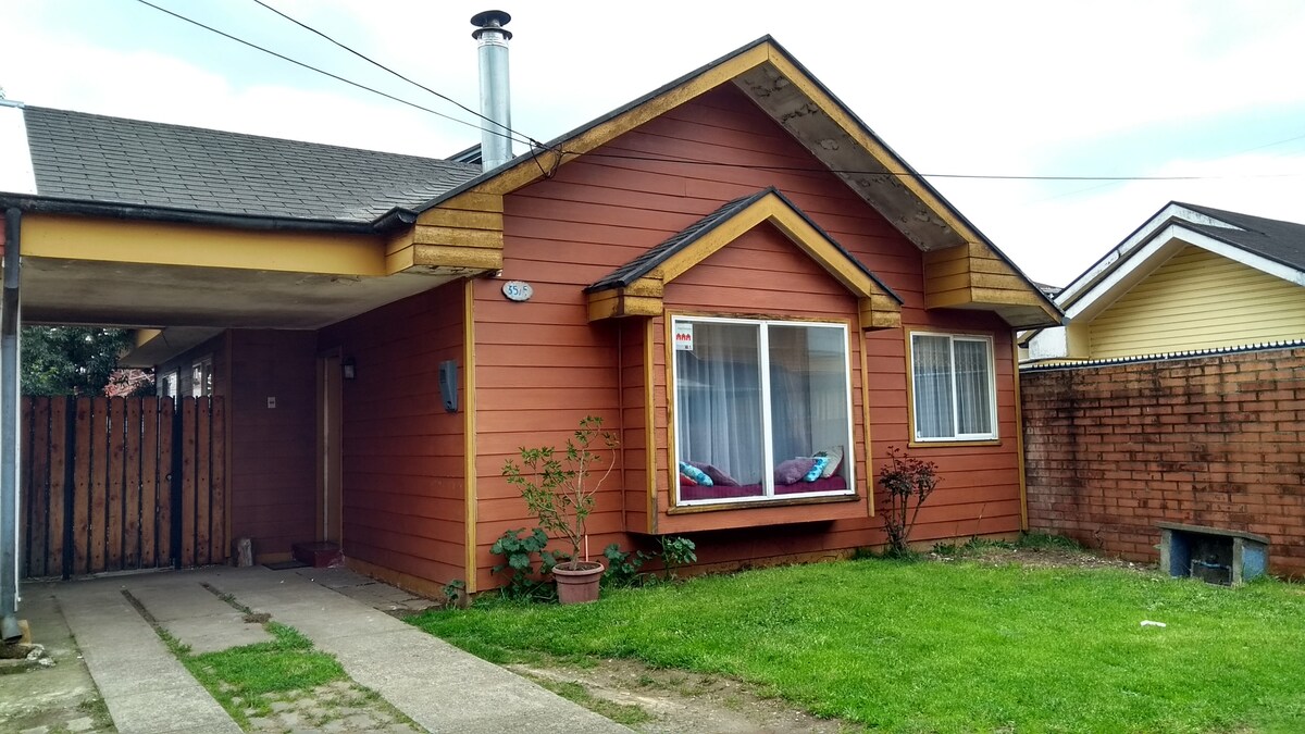 A single-story house features a warm brown exterior with a peaked roof. A spacious front yard displays green grass and decorative plants. Large windows allow natural light to enter, while a covered parking area is visible on the left side, providing convenient access.