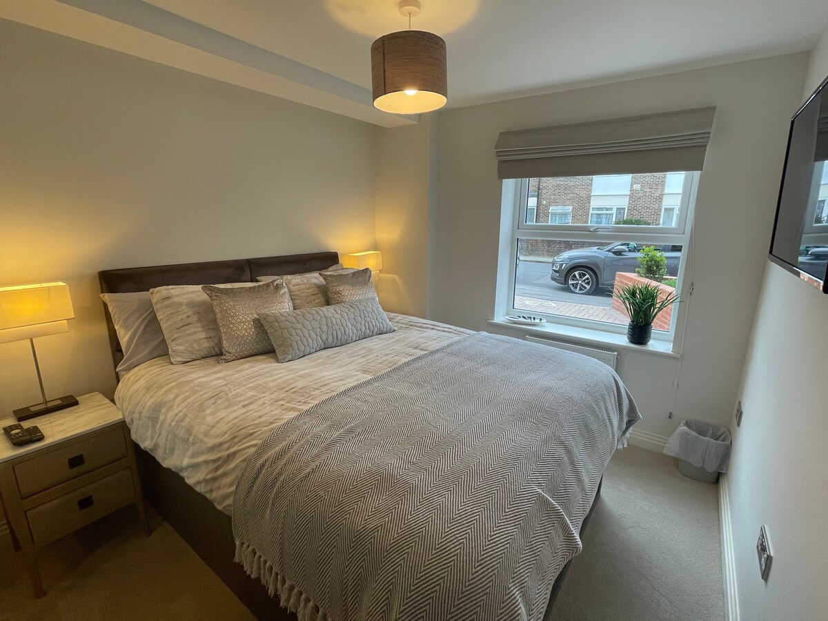 Dhomë gjumi e bukur dopio me televizor të montuar në mur, gardërobë të pajisur, grila të errëta dhe kabniet të ndriçuara pranë krevatit 