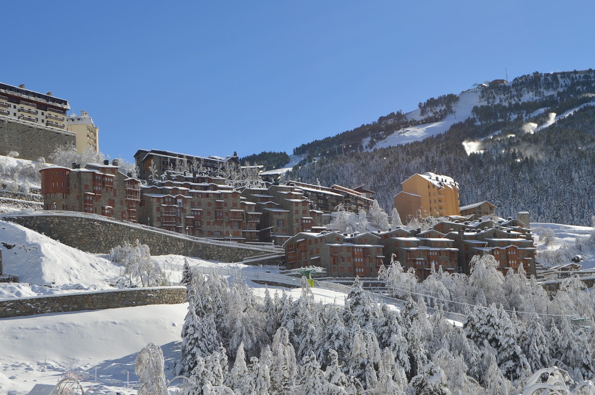 The image captures a snowy landscape featuring residential buildings nestled against a hillside. The architecture reflects a mix of styles, with warm-toned facades contrasting the white snow. Pine trees dusted with snow enhance the winter ambiance, framed by a clear blue sky.