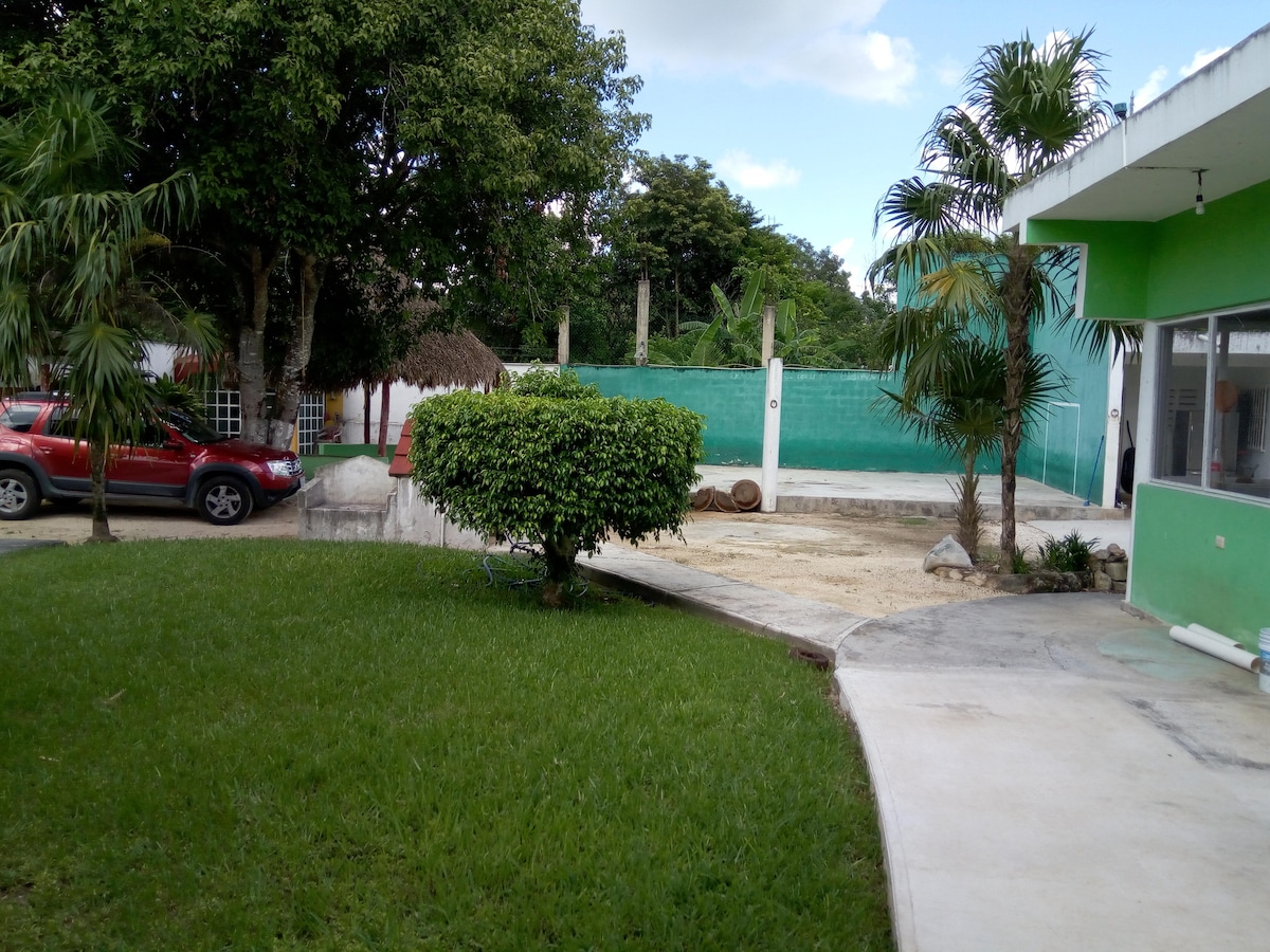 A grassy area is visible, bordered by tall palm trees. A vehicle is parked in a concrete area, alongside a well-maintained garden featuring a bush. In the background, a smooth surface and a palapa structure are also present, providing an inviting outdoor setting.
