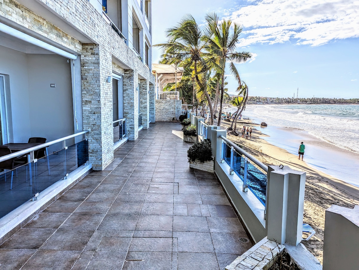 A spacious patio extends along the beachfront, featuring smooth stone flooring and a railing overlooking the ocean. Palm trees line the edge, providing a natural border. The sandy shore is visible in the background, with gentle waves lapping at the beach.