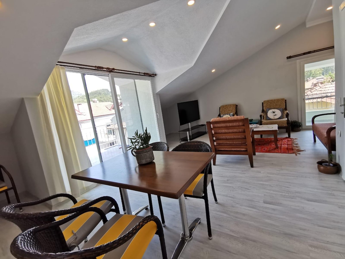 A light-filled living space features a sleek wooden table surrounded by four chairs. A comfortable seating area is present, facing a large window with views of the surroundings. A flat-screen television is mounted on the wall, and a small potted plant adds a touch of greenery.
