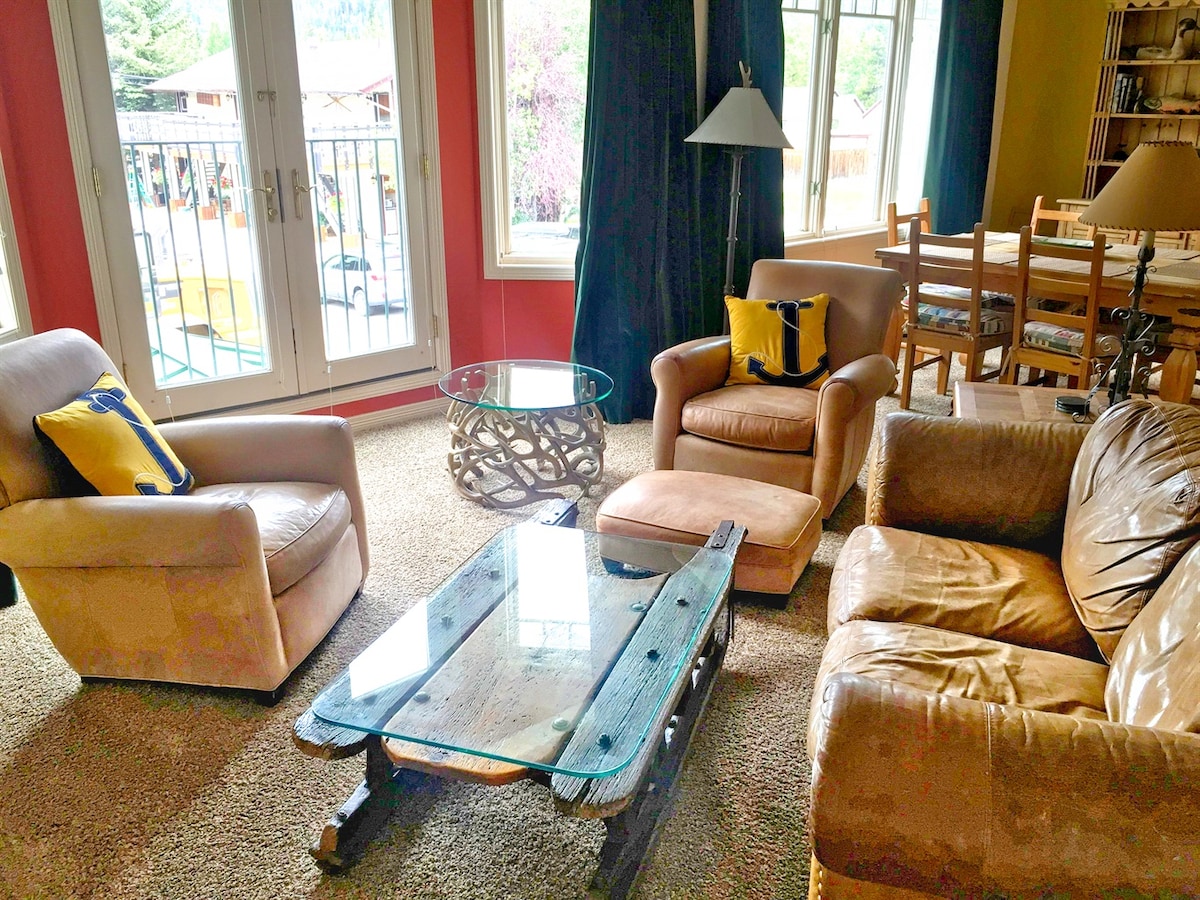 A cozy living area is displayed, featuring two comfortable armchairs and a leather sofa. A glass-topped coffee table rests on a carpeted floor. Large windows allow natural light to enter, with views of the outdoors and dark curtains providing contrast.